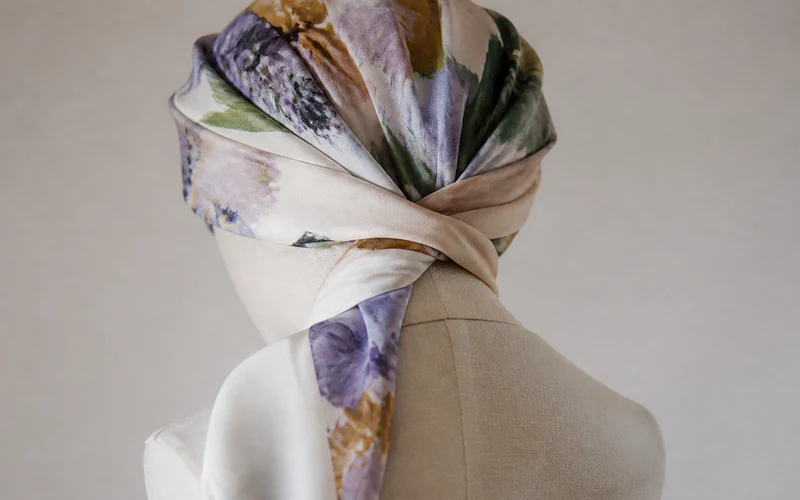 Mannequin head mid-tie showing a large square tichel being crossed at the nape with tails brought forward