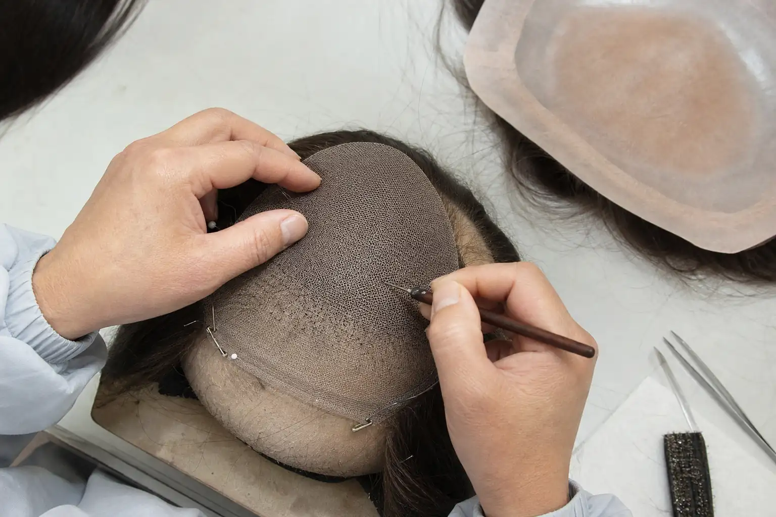 Illustrative close-up of an artisan ventilating dark hair into the lower crown mesh during production